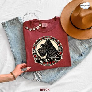 May include: Brick-colored t-shirt featuring a black Scottie dog illustration within a circular design with a plaid border and the text "scottie life." The shirt is paired with blue jeans, a brown hat, and a floral necklace.