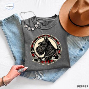 May include: A gray t-shirt featuring a black Scottish Terrier dog illustration within a circular design with red plaid accents. The text "scottie life" is displayed on a banner below the dog. A brown hat and a floral necklace are also visible.