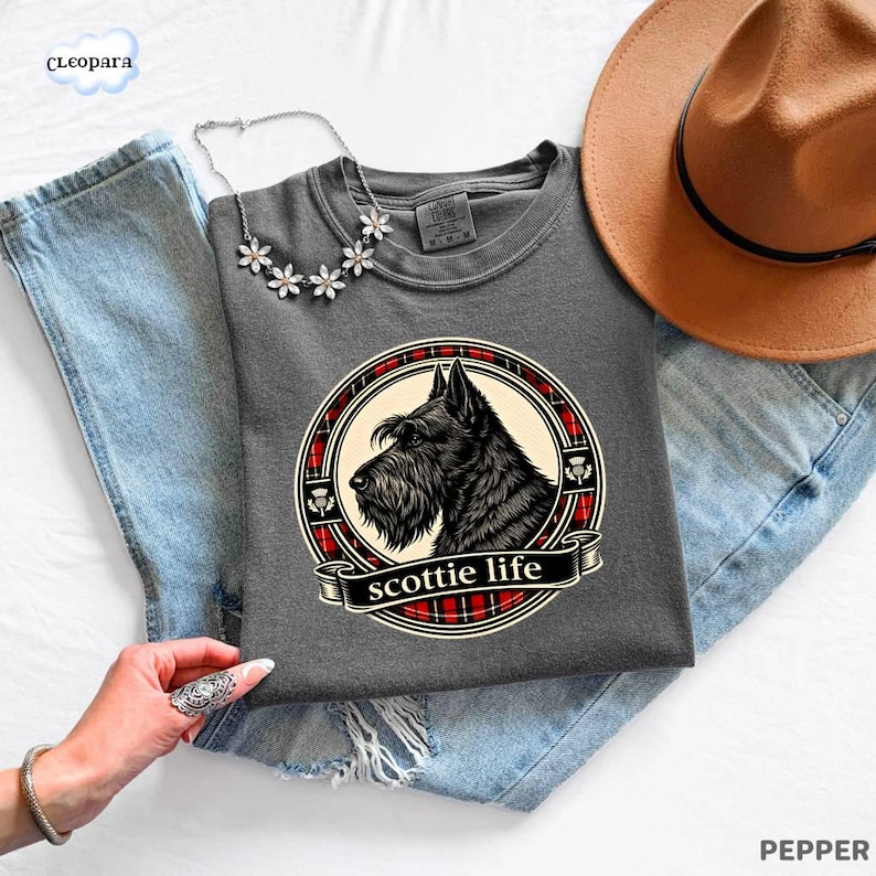 May include: A gray t-shirt featuring a black Scottish Terrier dog illustration within a circular design with red plaid accents. The text "scottie life" is displayed on a banner below the dog. A brown hat and a floral necklace are also visible.