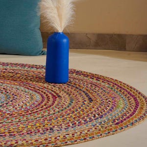 May include: A round, multi-coloured braided jute rug featuring concentric circles of various hues. Behind the rug, a brown square cushion with a geometric pattern sits next to a solid teal cushion. A bright blue vase holds white pampas grass.