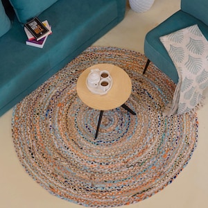 Può includere: Un tappeto rotondo intrecciato multicolore con un tavolino in legno e un divano e una sedia turchese. Il tappeto presenta sfumature di beige, arancione, blu e viola. Una coperta bianca con un motivo a foglie è drappeggiata sulla sedia.
