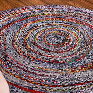 May include: A round braided rug constructed from multicolored fabric strips. The design features concentric circles of woven material in shades of blue, grey, red, yellow, and green, creating a textured, multi-tonal surface pattern throughout the piece.