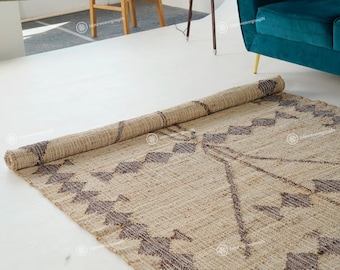 May include: A rolled-up, beige jute rug with a geometric pattern in dark brown. The rug is partially unrolled, revealing the design. The rug is on a white surface, with a teal sofa and wooden chair in the background.