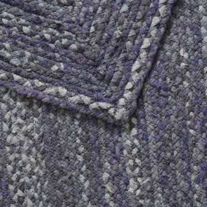 May include: Close-up of a braided rug with a mix of gray, white, and purple hues. The rug's texture is visible, with a detailed pattern of interwoven strands. The corner of the rug is folded over, showing the edge.