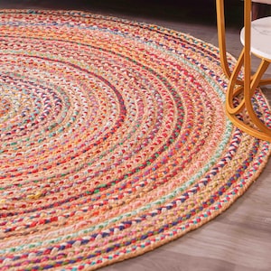May include: A round, braided rug with a spiral design. The rug features a mix of colors, including orange, pink, blue, and green. The rug is made of a natural fiber and has a textured appearance. The rug is placed on a wooden floor.