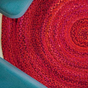 Puede incluir: Una alfombra redonda trenzada en tonos rojos y rosas. El diseño de la alfombra presenta un patrón en espiral, con el centro en un tono más oscuro de rojo. La alfombra está hecha de un material tejido y tiene una superficie texturizada.
