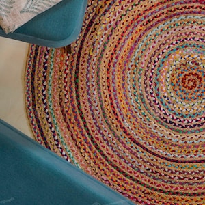 Puede incluir: Una alfombra redonda de yute trenzado con un diseño en espiral. La alfombra presenta una mezcla de colores, incluyendo marrón, naranja, rojo, azul y morado. La alfombra está junto a una silla turquesa.