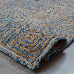 May include: A rectangular area rug crafted from braided natural jute and blue denim fabric strips. The rug features a repeating geometric square pattern throughout, with one corner folded over to reveal the thick, textured woven construction.