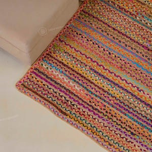 Op de afbeelding: Een rechthoekig, veelkleurig gevlochten jute vloerkleed met een roze rand. Het vloerkleed heeft strepen in verschillende kleuren, waaronder oranje, blauw, paars en groen. Het vloerkleed ligt op een wit oppervlak.