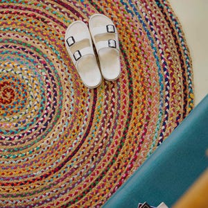 Könnte beinhalten: Ein runder, mehrfarbiger geflochtener Jute-Teppich mit Spiralmuster. Zwei weiße Sandalen mit schwarzen Schnallen liegen auf dem Teppich. Der Teppich hat Beige-, Rot-, Gelb-, Blau- und Violetttöne. Im Hintergrund ist eine türkisfarbene Fläche.