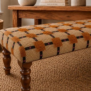 May include: A rectangular upholstered bench featuring a textured beige fabric base with a grid pattern of raised orange squares, thin black lines, and small pink accents. The bench is supported by four dark brown turned wooden legs.