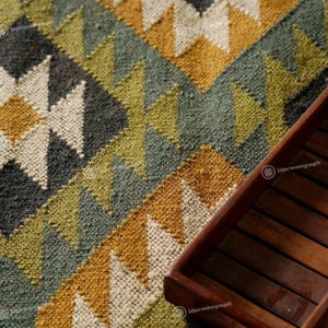 May include: Close-up of a hand-woven rug with a geometric pattern in shades of gray, white, yellow, and green. A dark brown wooden tray sits on the right side of the rug. The rug's design features diamond and star-like shapes.