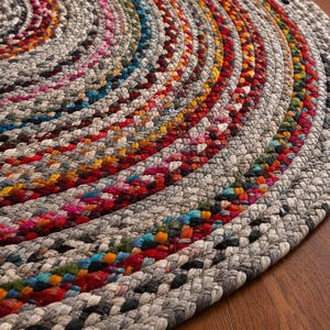 Puede incluir: Una alfombra redonda trenzada que presenta círculos concéntricos de cuerda gruesa y texturizada. El diseño incorpora un color base gris neutro intercalado con hebras vibrantes y multicolores de rojo, amarillo, azul y rosa en el tejido.