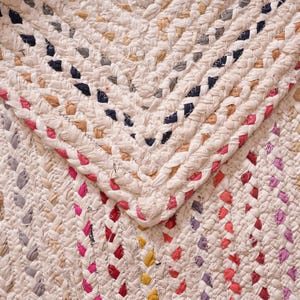 May include: Close-up of a braided rug with a cream-colored base and colorful accents. The rug features a chevron pattern with red, pink, blue, and yellow details. The braided texture adds visual interest to this home decor item.