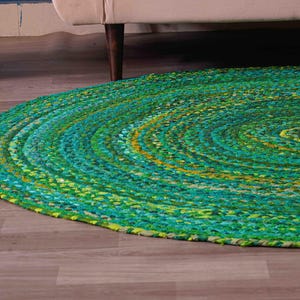 Könnte beinhalten: Ein runder geflochtener Teppich in Grün-, Türkis- und Gelbtönen. Der Teppich besteht aus einem dicken, strukturierten Material und hat ein Spiraldesign. Er liegt auf einem Holzboden, mit einem weißen Korb und dem Bein eines beigen Sofas im Hintergrund.