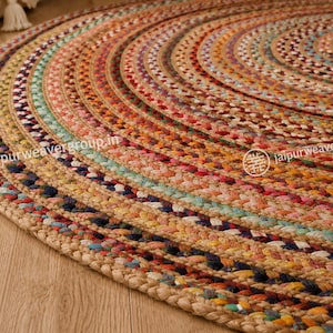 May include: A round braided rug featuring concentric circles of multi-colored fabric strips in shades of red, blue, yellow, green, and natural tan jute. The surface displays a textured, woven pattern with the text jaipurweavergroup.in visible.