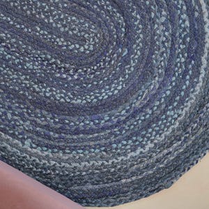 May include: Oval braided rug in shades of blue and gray. The rug has a spiral pattern and is made of a woven material. The rug is a home decor item.