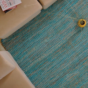 May include: A close-up shot of a turquoise and tan woven rug. The rug is rectangular and has a textured appearance. A yellow decorative object is on the rug. Beige furniture is in the background, with a book on top.