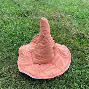 May include: A quilted peach-colored pointed hat with a wide, circular brim. The fabric features a diamond-shaped stitched pattern across the entire surface, including the tall, conical crown and the flat, rounded brim edge.