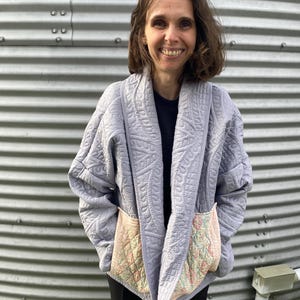 May include: A light gray quilted jacket with large pockets. The jacket has a shawl collar and a quilted pattern. The pockets are a contrasting peach color with a floral pattern. The jacket is worn over a black shirt.