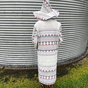 May include: A long, quilted coat and a matching pointed hat, both featuring a cream-colored base with horizontal bands of intricate geometric patterns in shades of blue, red, and grey. The coat reaches mid-calf and has long sleeves.