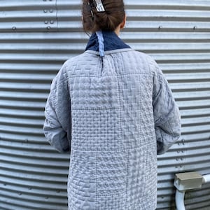 May include: A light gray quilted jacket with a navy blue collar and light blue trim. The jacket features intricate stitching patterns and is worn by a person with a hair clip. The background is a corrugated metal surface.
