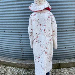May include: A white quilted coat and matching hat, both adorned with a delicate floral pattern in shades of pink and green. The coat is long, reaching the ankles, and the hat has a wide brim with a pink trim. The outfit is worn outdoors.