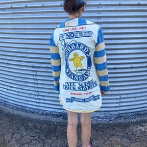 May include: A long-sleeved jacket constructed from repurposed feed sack fabric featuring a cream-colored back panel with a graphic of a yellow chick and the text Hubbard's Standard All Mash Chick Starter. The sleeves have blue and yellow stripes.