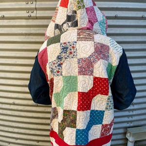 May include: A hooded patchwork jacket with a quilted design. The jacket features a variety of colorful fabric squares in red, blue, green, and floral patterns. The hood and hem are trimmed in red. The sleeves are a dark blue.