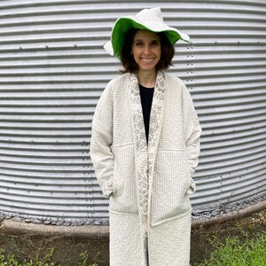 May include: A long, quilted coat in a light beige color with a subtle white striped pattern and a wide, floral-patterned lapel. The coat features long sleeves and deep pockets. A matching wide-brimmed hat with a bright green underside is worn.