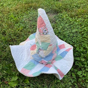 May include: A conical hat constructed from quilted fabric patches in pastel shades of pink, blue, green, and white. The hat features a wide, floppy brim and a tall, pointed crown, with a patchwork design consisting of floral prints and polka dots.