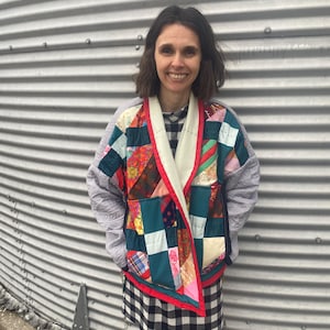 May include: A patchwork jacket with a variety of colorful fabric squares, including red, green, and blue, is worn over a black and white checkered dress. The jacket has a white collar and red trim.