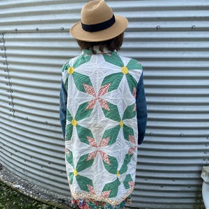 May include: A tan straw fedora with a black band worn with a long jacket featuring a white, green, and yellow star quilt pattern on the back. The jacket has dark blue denim sleeves and a floral print hem, worn over a colorful floral patterned skirt.