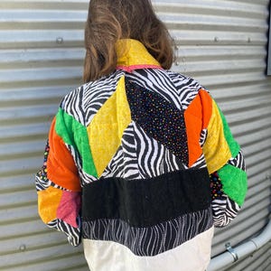 May include: A colorful patchwork jacket with a variety of patterns and colors. The jacket features zebra print, black polka dots, and solid blocks of orange, yellow, green, and black. The bottom hem is white with a pink trim.