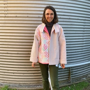 May include: A light pink quilted jacket with a patchwork design on the front. The jacket has a collar and is worn over a gray turtleneck and olive green pants. The jacket's interior is a bright pink color.