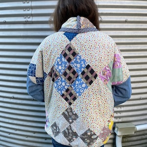 May include: A quilted jacket with a patchwork design. The jacket features a variety of fabric patterns, including floral prints, plaid, and solid colors. The sleeves are denim. The jacket is worn by a person.