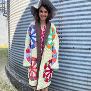 May include: A long-sleeved quilted coat featuring a cream-colored base with large, circular, multi-colored patchwork patterns in red, blue, orange, and green. The coat has a dark brown collar and trim, paired with a wide-brimmed, light-colored hat.