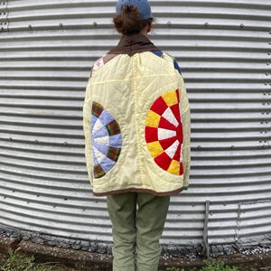 May include: A quilted jacket featuring a pale yellow base with large circular fan-shaped patterns in red, white, and blue. The jacket has a dark brown collar, long sleeves, and a boxy fit, constructed from patchwork fabric with visible stitching.