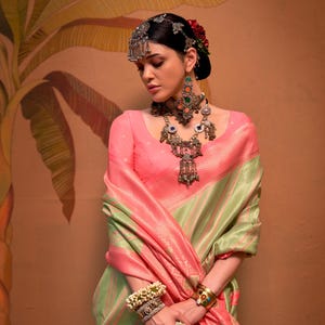 May include: A woman wearing a pink and green sari with intricate jewelry. The sari is draped over the shoulders and arms. The jewelry includes a headpiece, earrings, a necklace, bracelets, and rings. The background is a warm, earthy tone.