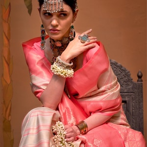 May include: A woman wearing a pink and cream sari with intricate jewelry, including a headpiece, earrings, necklace, ring, and bracelets. The sari has a detailed pattern and is draped elegantly. The overall aesthetic is traditional and ornate.