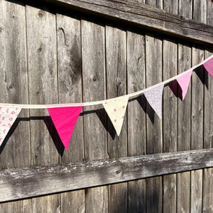 Peut inclure: Une guirlande de fanions en tissu colorés, roses et blancs, avec des motifs floraux et à pois. Les drapeaux triangulaires sont enfilés sur un ruban blanc, créant une décoration festive. L'arrière-plan est une clôture en bois vieilli.