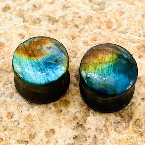 May include: Two round ear plugs with a dark base and a colorful, iridescent top. The tops feature shades of blue, green, and gold, creating a marbled effect. The plugs are displayed on a textured, light brown surface.