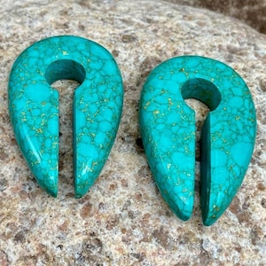 May include: Pair of turquoise teardrop-shaped ear gauges with a split design. The gauges have a polished finish and a natural stone appearance with gold-colored veining. The teardrops are displayed on a textured, light brown surface.