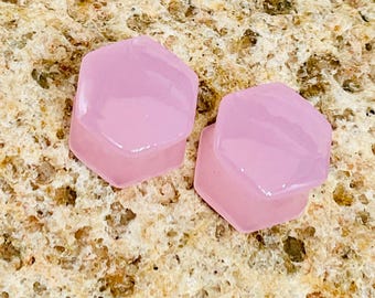 Pode incluir: Dois plugues hexagonais rosa claro. Os plugues são feitos de um material translúcido e têm uma superfície lisa e polida. Os plugues são exibidos em uma superfície texturizada e marrom.