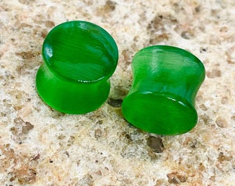 May include: Two bright green ear plugs. The plugs have a rounded, flared shape with a smooth, glossy finish. The plugs are made of a solid, opaque material and are designed to fit into stretched ear piercings. The plugs are displayed on a textured surface.