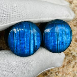Peut inclure: Une paire de bouchons d'oreille ronds bleus avec un motif rayé. Les bouchons ont une finition brillante et sont tenus par un tissu blanc. La couleur bleue a un effet chatoyant, et les bouchons semblent être en pierre.