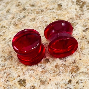 May include: Two translucent red ear gauges. The plugs have a flared design with a rounded front and back. The gauges are made of a glossy material and are displayed on a textured, light brown surface.