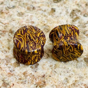 May include: Two wooden ear plugs with a unique pattern. The plugs are cylindrical with flared ends. The pattern is a mix of yellow and brown, creating a textured appearance. The plugs are designed for stretched ear piercings.
