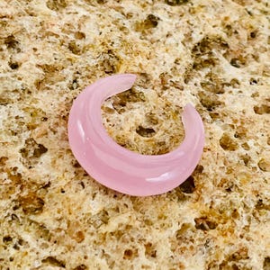 May include: A pale pink, crescent-shaped earring. The earring is made of a translucent material and has a smooth, curved design. The earring is set against a textured, tan background.