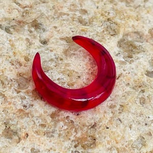 Könnte beinhalten: Ein leuchtend roter, halbmondförmiger Ohrring. Der Ohrring besteht aus einem durchscheinenden Material mit dunkleren roten Marmorierungen. Der Ohrring liegt auf einer strukturierten, hellbraunen Oberfläche.